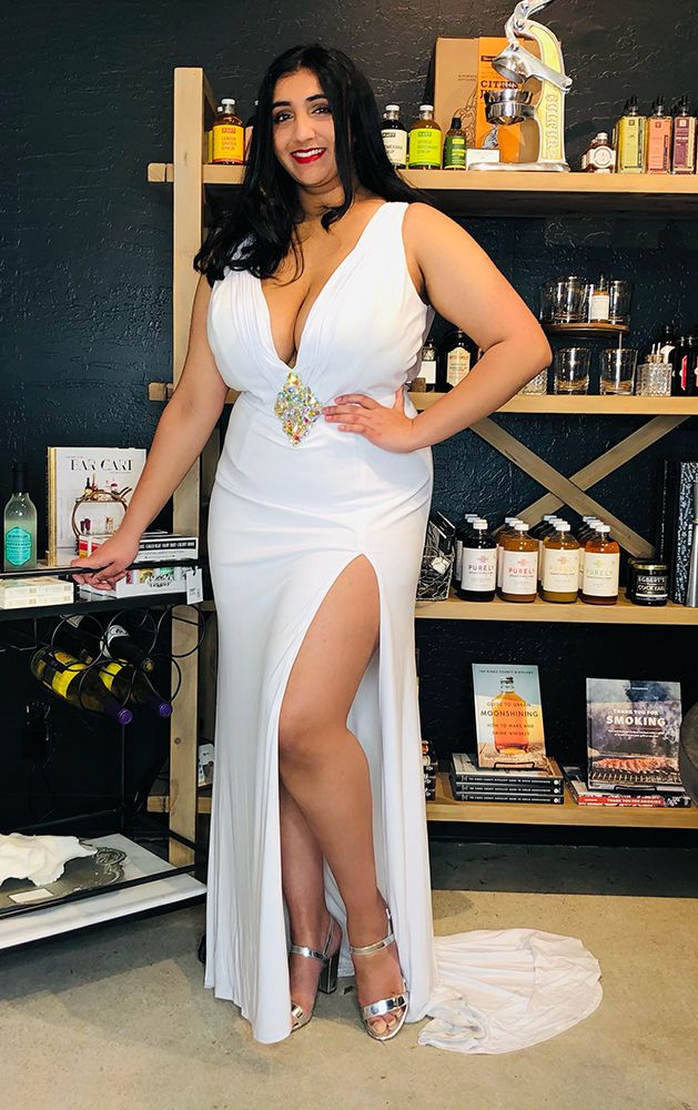 model in form-fitting white sleeveless gown with plunging neckline, high slit, diamond-shaped rhinestone embellishment at waist, and silver chunky-heel sandals