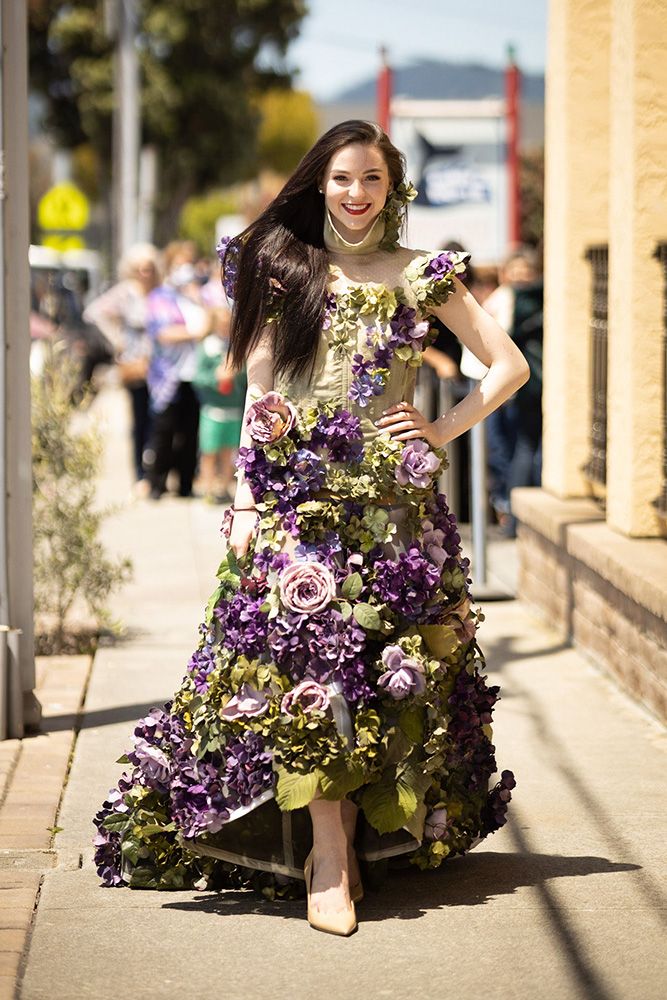 smiling model in a long light green gown covered with pink and purple 3-D flowers