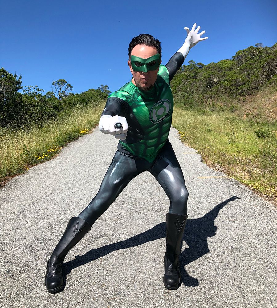 model in action pose while wearing gray and green unitard with printed-on abs and highlights, and green mask in a coastal trail setting