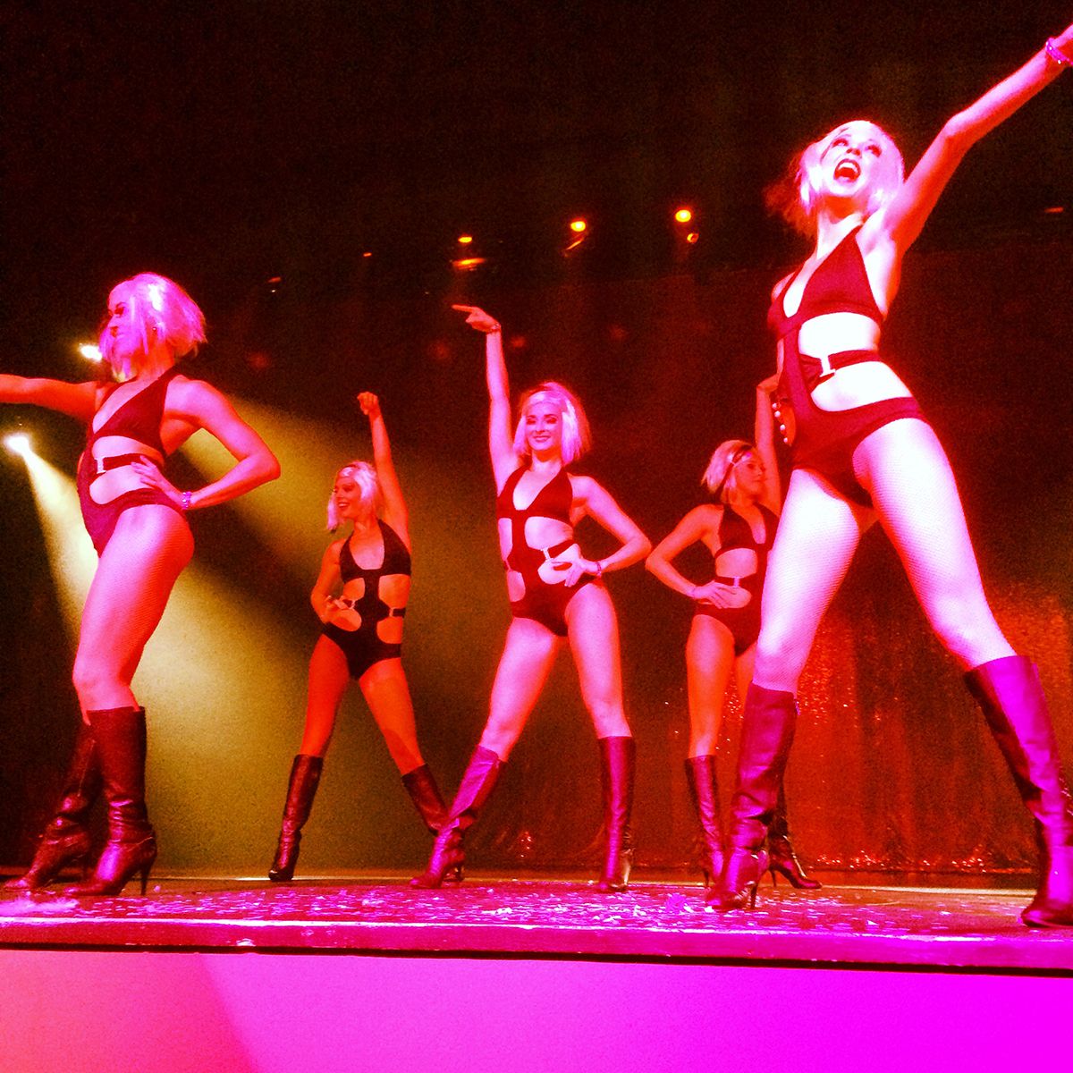 five girls on stage in black keyhole bodysuits, arms raised, bathed in pink light
