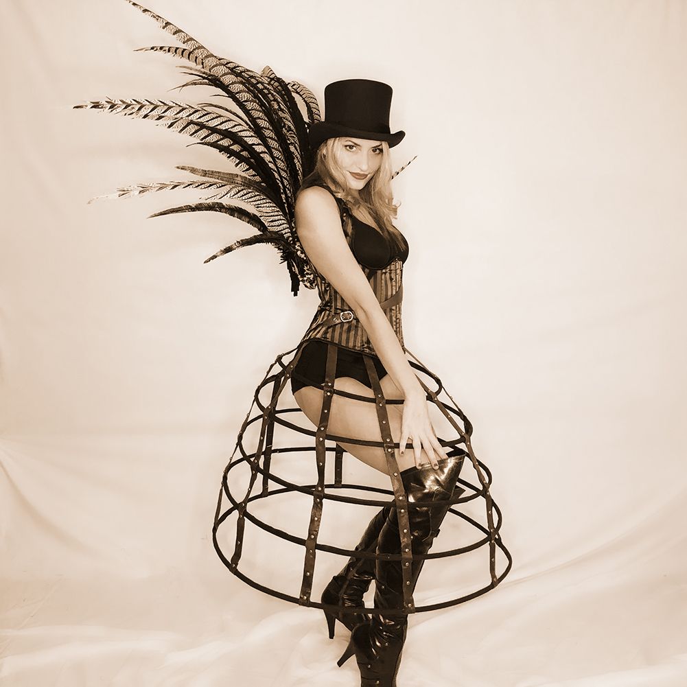 sepia-toned photo of woman in body suit and knee-high boots as well as pheasant feather back piece, leather hoop skirt staves, top hat, and corset
